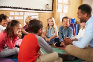 A teacher and young students sit in a circle on a classroom rug, smiling and talking together near a whiteboard and colorful storage shelves.